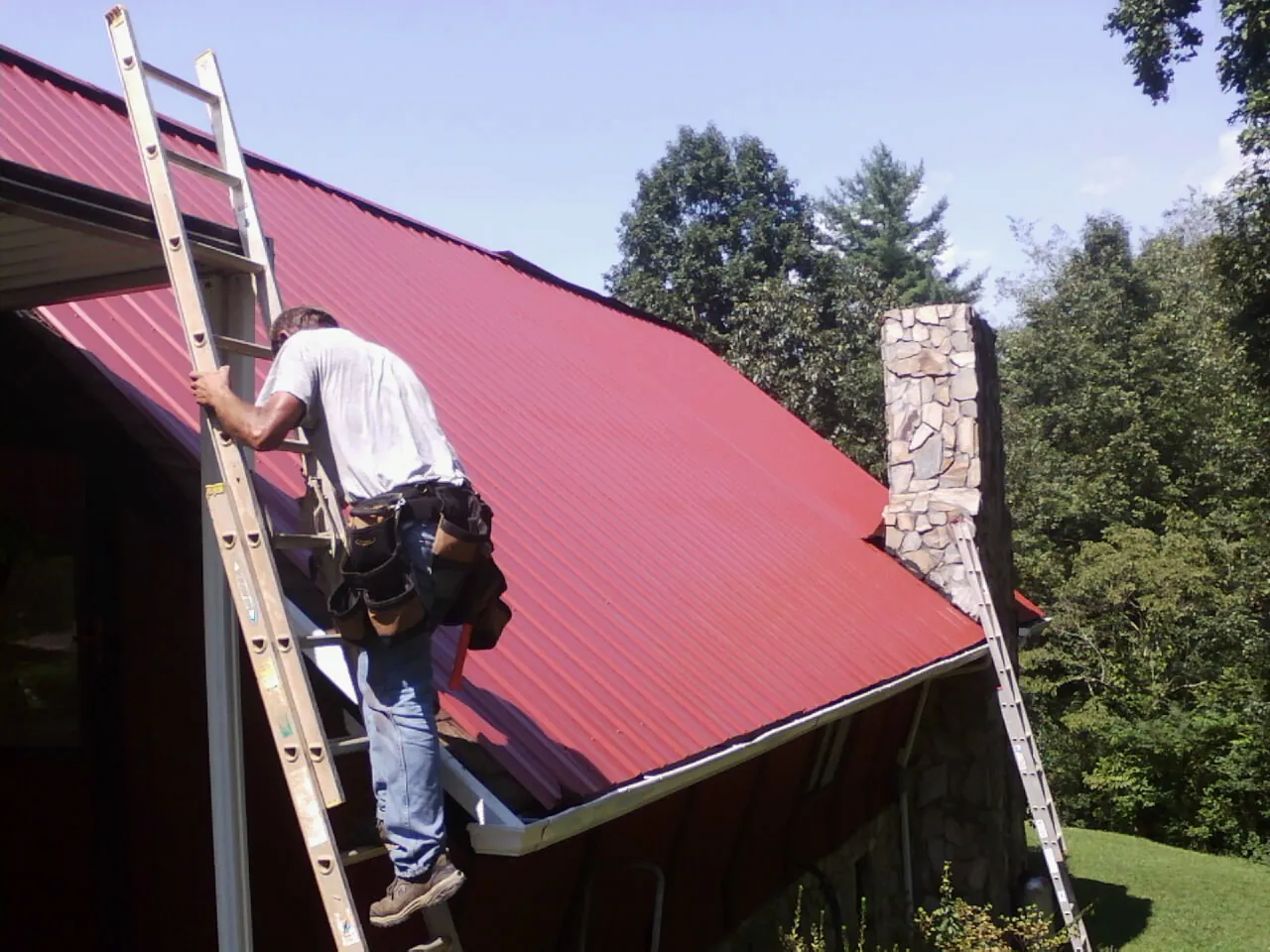 Roofer on a ladder beside a red metal roof and stone chimney — Soffit & Fascia Repair by Roofing Brothers in Winfield